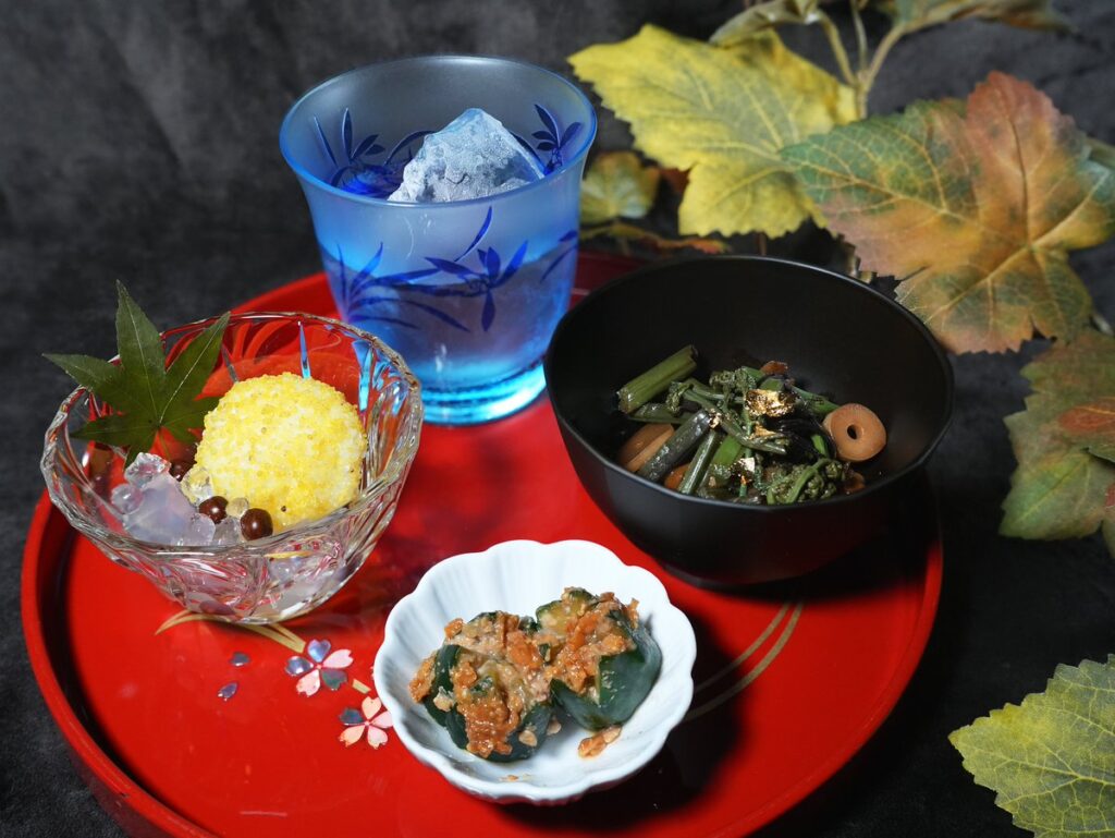 朱塗りの盆に載せた『遠野物語』日本酒セット。青いグラスの冷酒に胡瓜の赤味噌漬け、山菜の油炒め、粟餅のみつ豆風