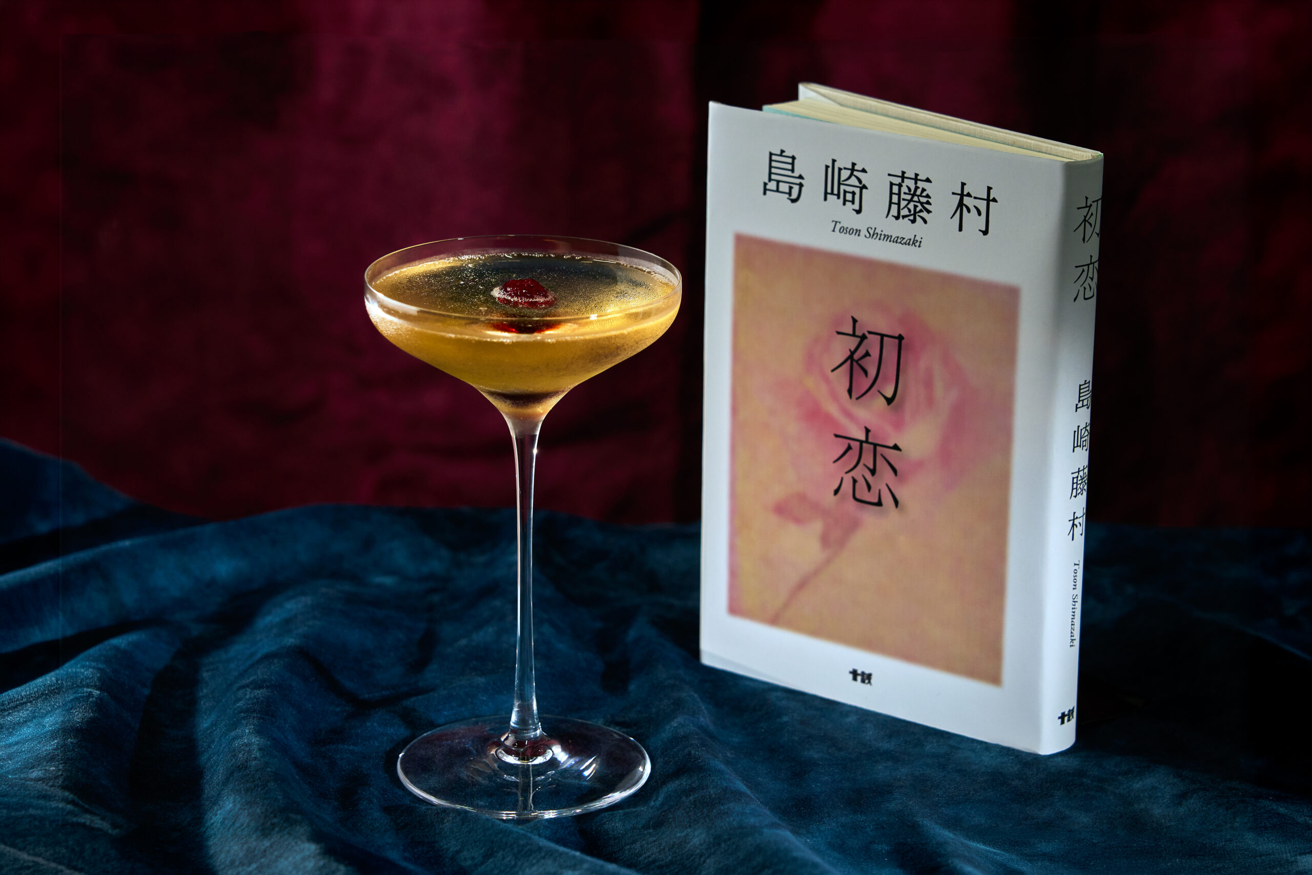 島崎藤村「初恋」をイメージした桑酒とシャンパンの金色のカクテル。クープグラスに赤い実を浮かべ、薔薇の表紙の本を添えて