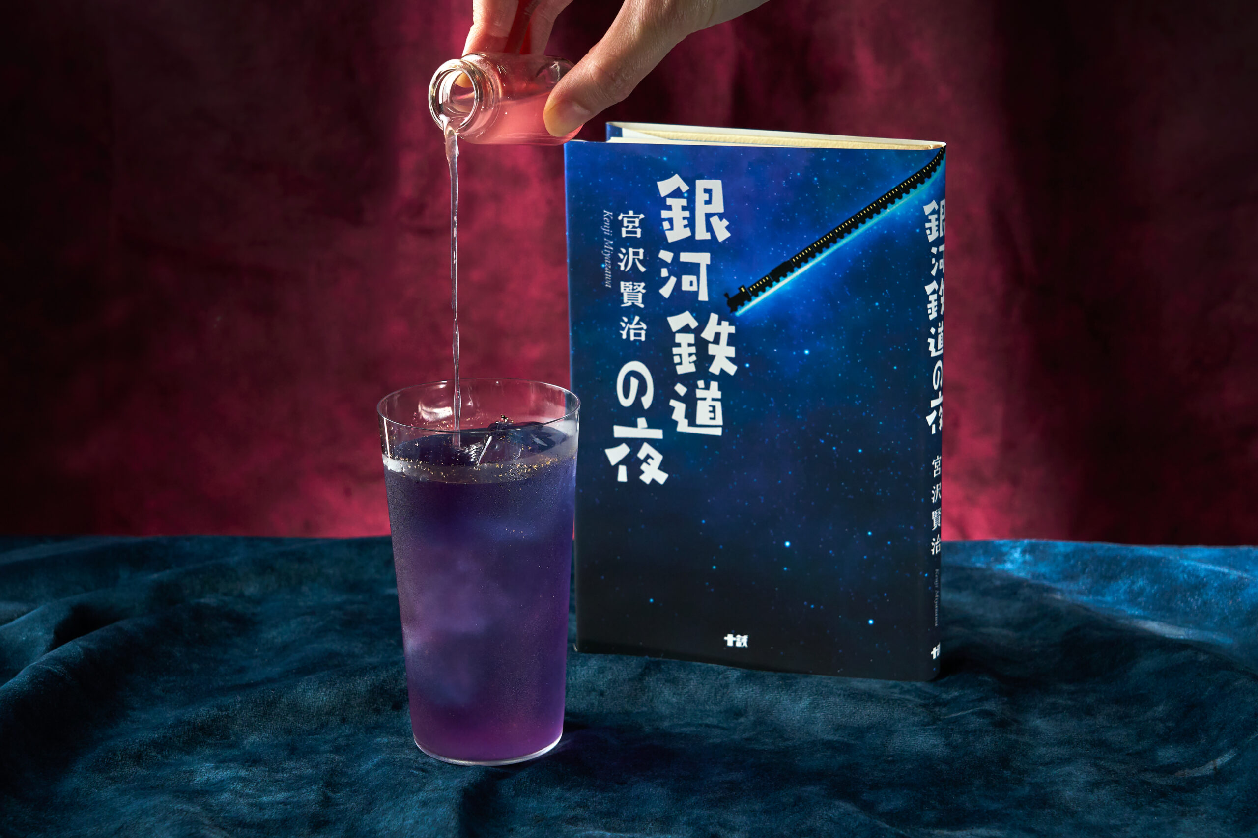 『銀河鉄道の夜』の本の隣で、金箔の星屑が浮かぶ青紫のギャラクシーハーバルレモネードに小瓶のシロップを注ぐ